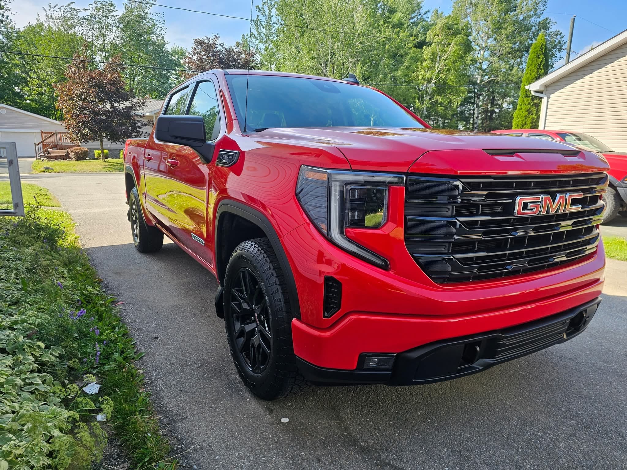 A bright red GMC pickup truck is parked in a driveway on a sunny day, surrounded by houses, trees, and greenery.