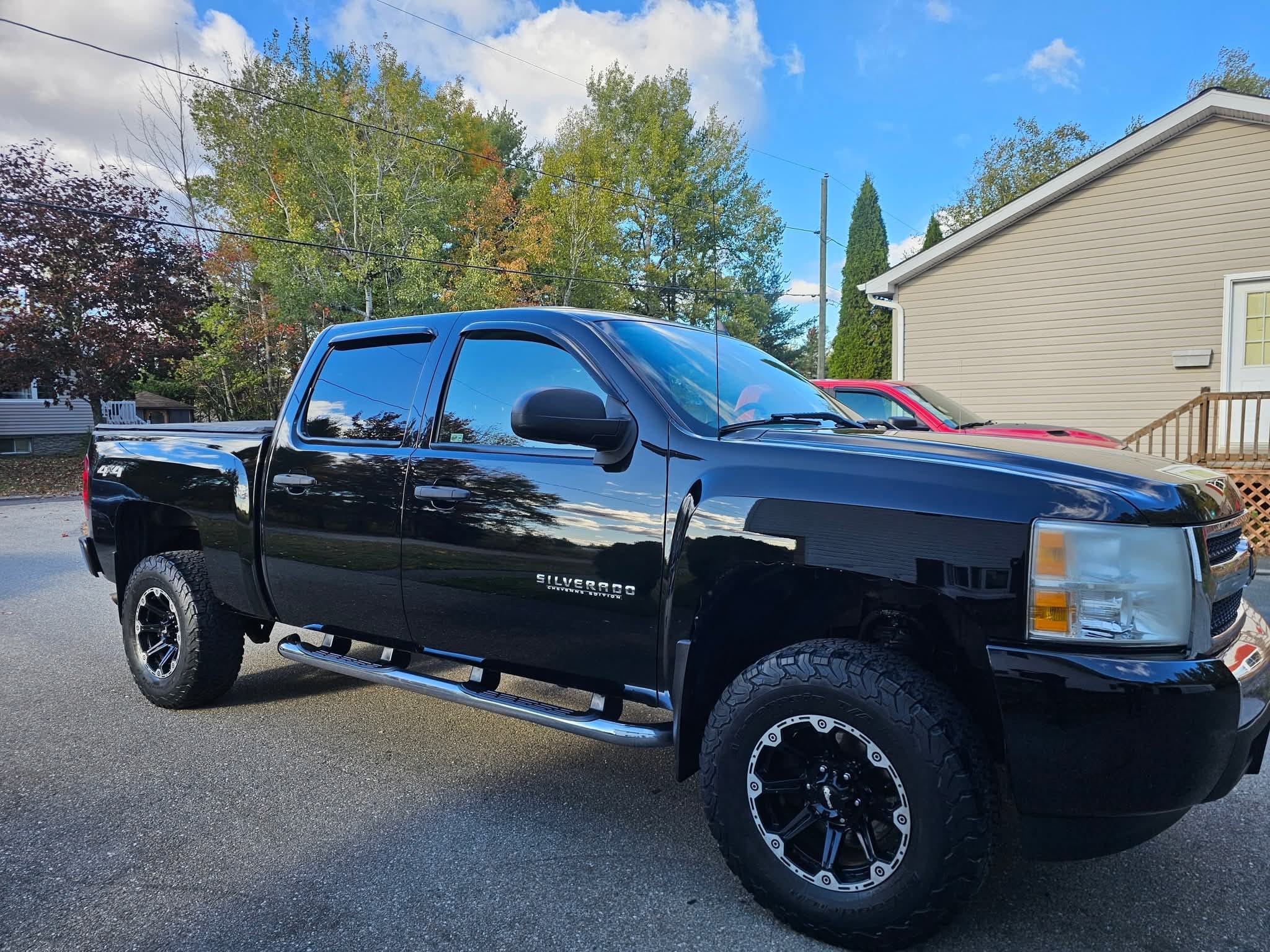 A black Chevrolet Silverado pickup truck with off-road tires is parked on a driveway in front of trees and a beige house on a sunny day.
