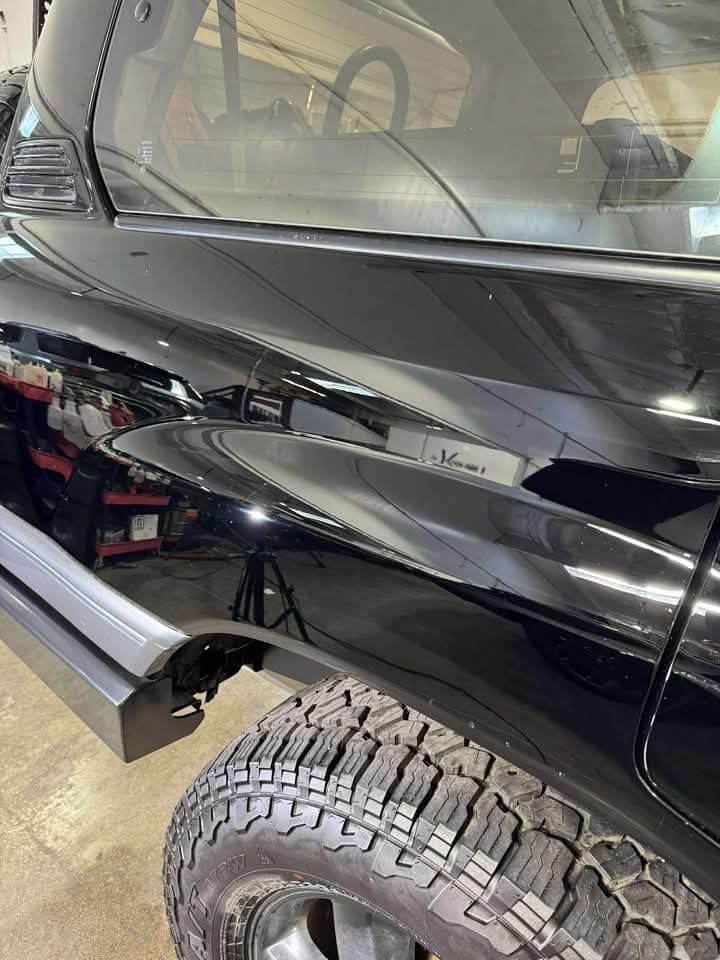 A close-up of the rear side of a shiny black vehicle, showing a reflection of the garage interior. The vehicles tire and part of the bumper are visible.