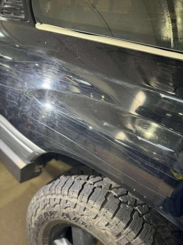 A close-up of a black vehicle shows scratches and a noticeable dent above the rear wheel, with a reflection of light highlighting the damage on the body.