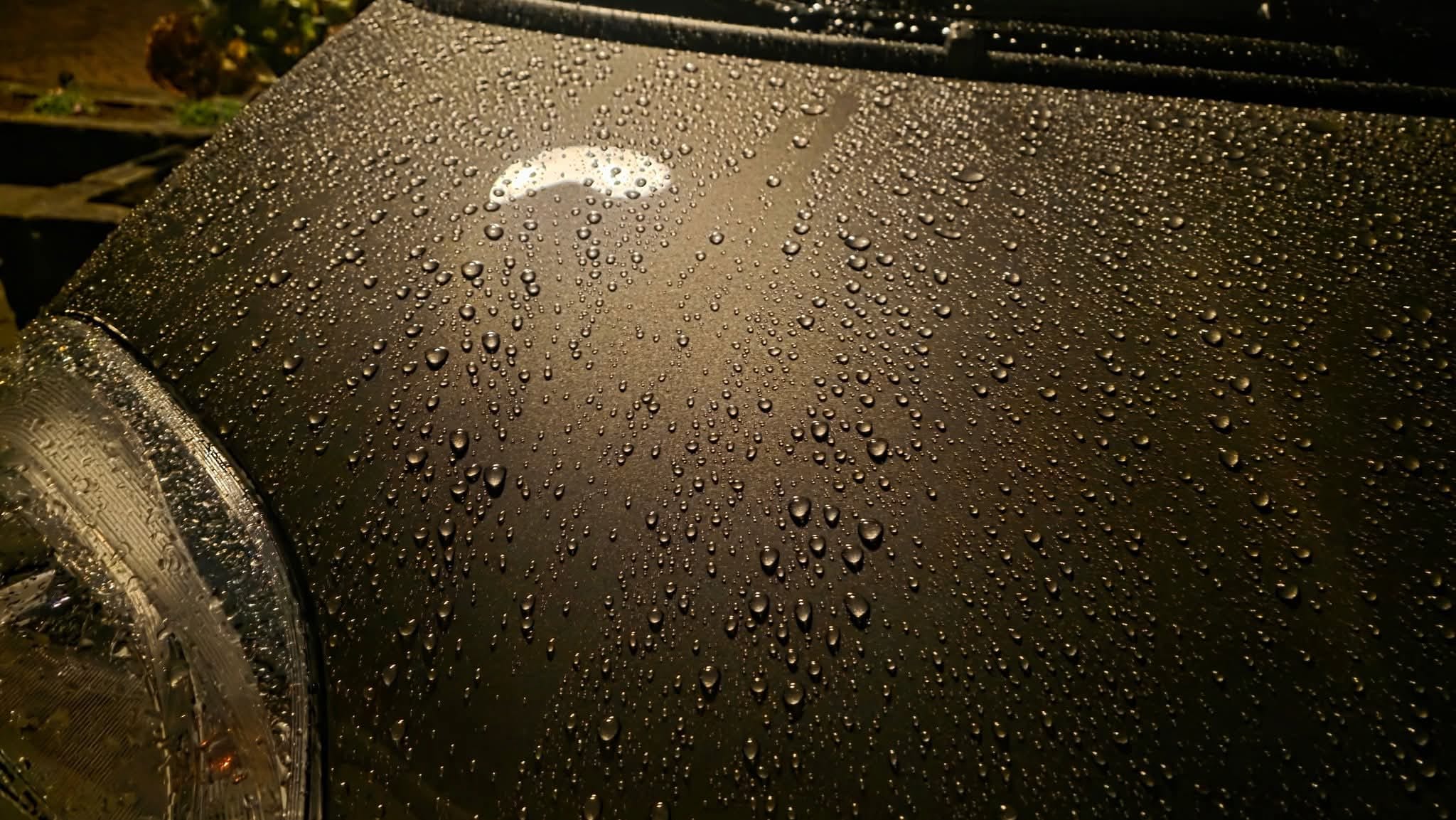 Close-up of a car hood covered in water droplets, reflecting warm yellow light. The surface is smooth and shiny, with a headlight partially visible in the lower left corner.