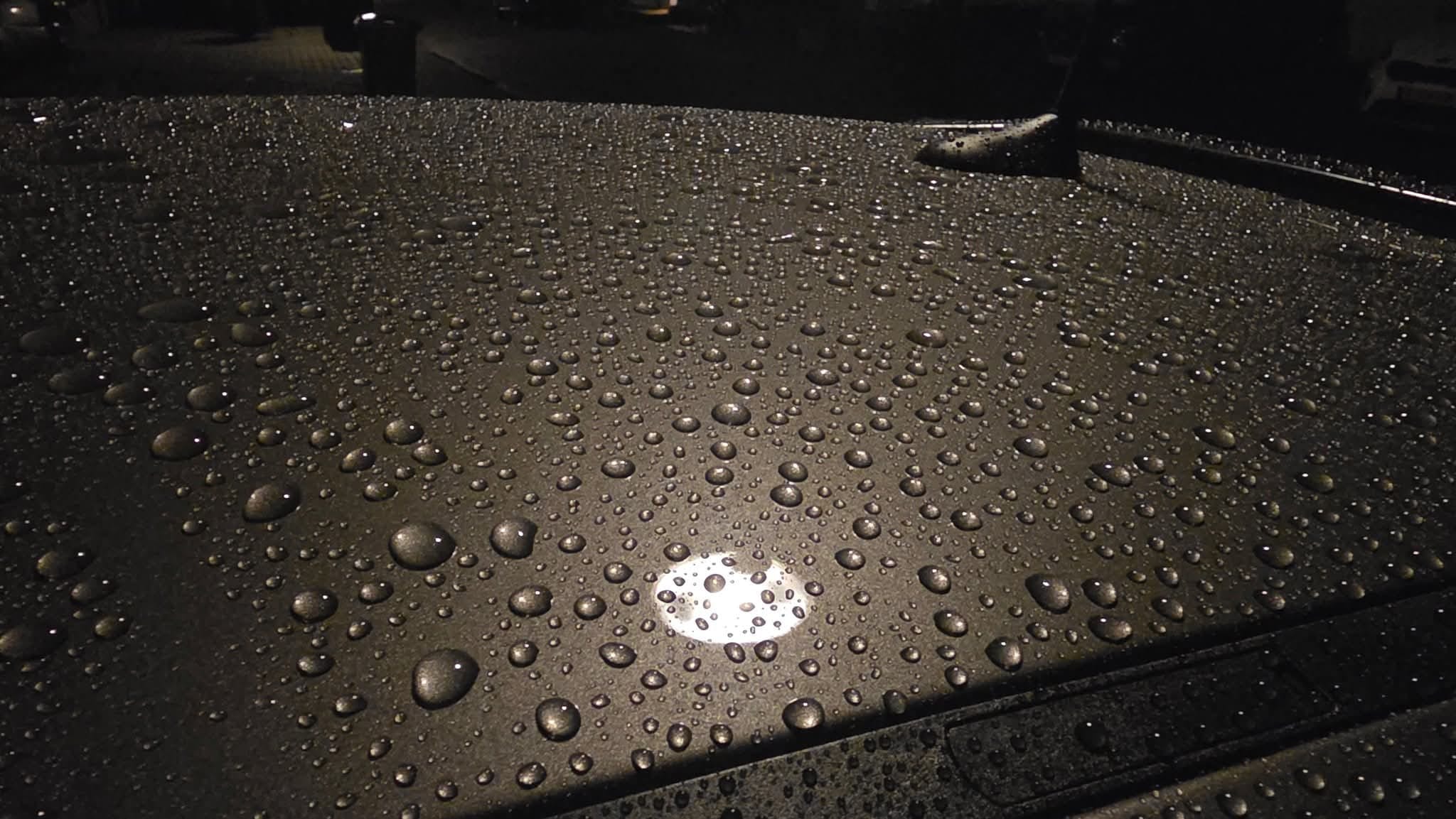 Raindrops scattered across the surface of a dark car roof at night, reflecting a bright circular light and some streetlight, creating a pattern of small and large water droplets.